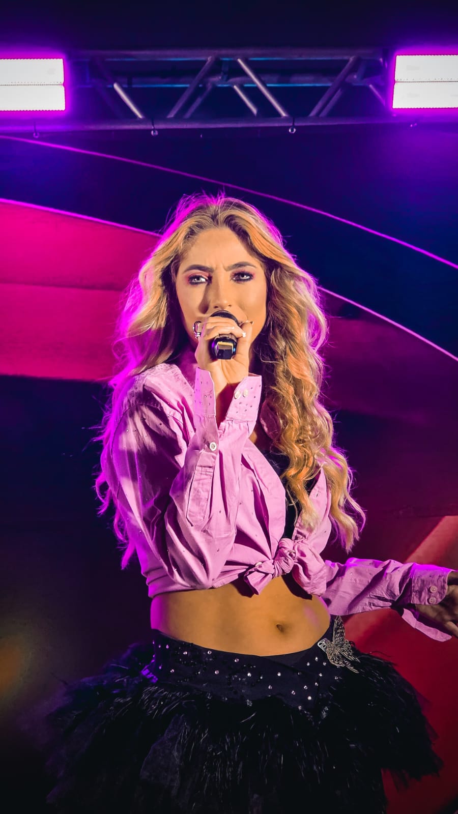 Jessy Portilla no palco, camisa rosa e saia de plumas preta, luz roxa