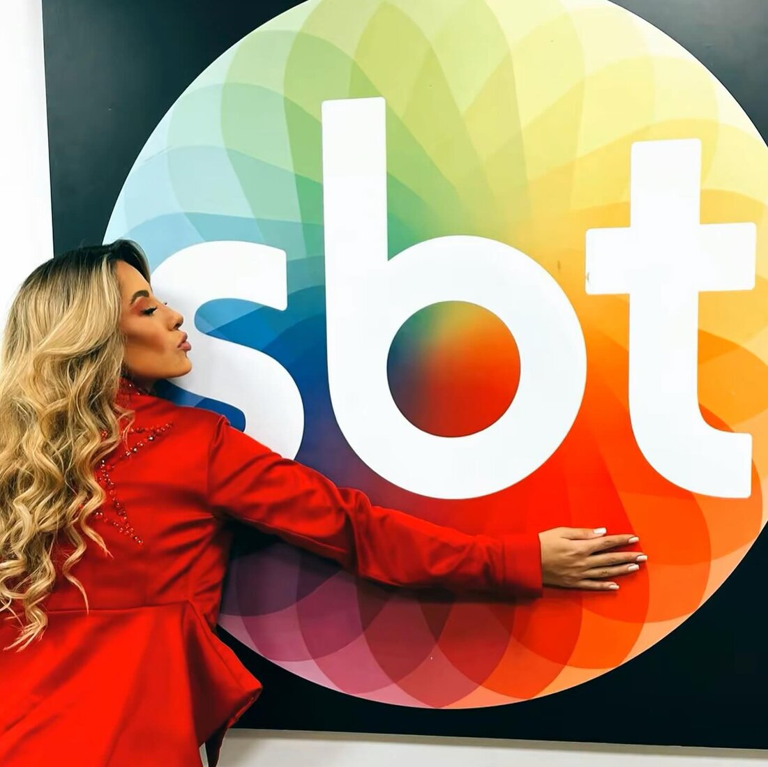 Jessy Portilla no estúdio do SBT, com blazer vermelho, abraçando o logo da emissora